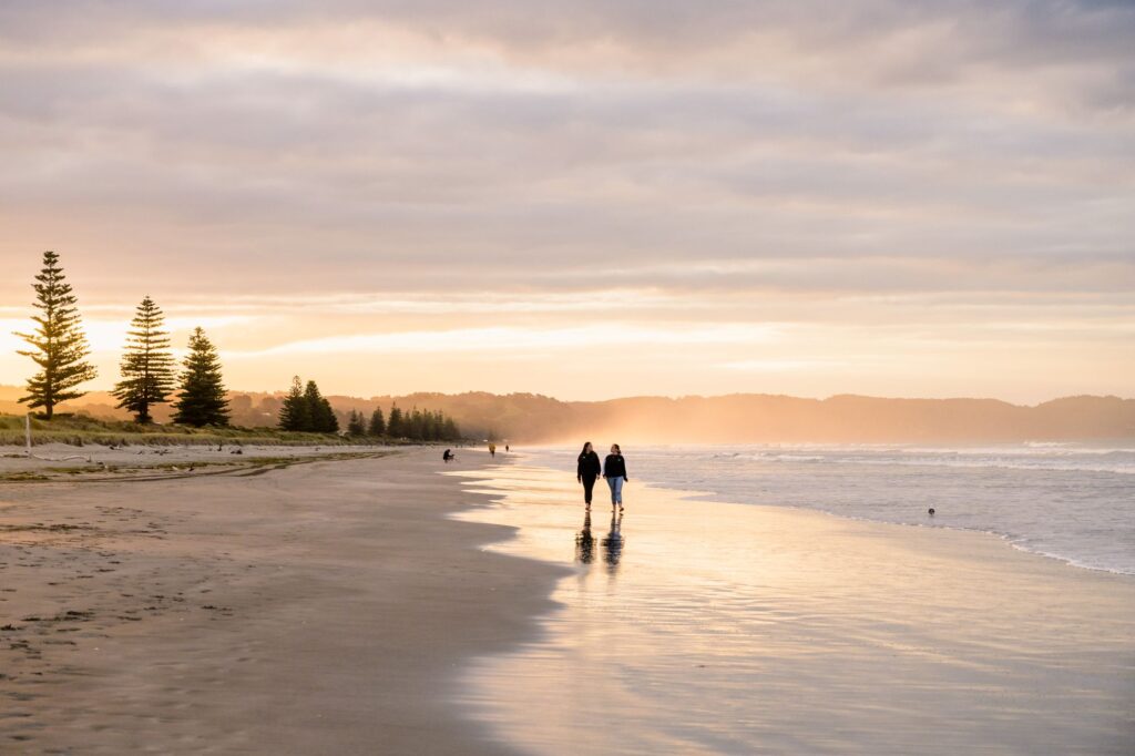 Two people walking along Ōhope Beach at sunset, with golden light reflecting off the wet sand and pine trees lining the shore.