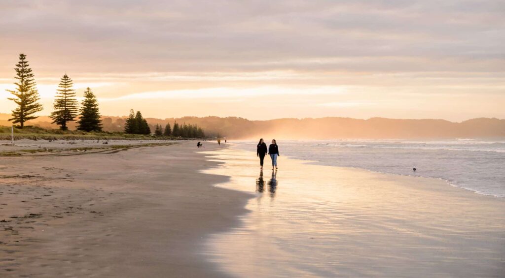 Two people walking along Ōhope Beach at sunset, with golden light reflecting off the wet sand and pine trees lining the shore.