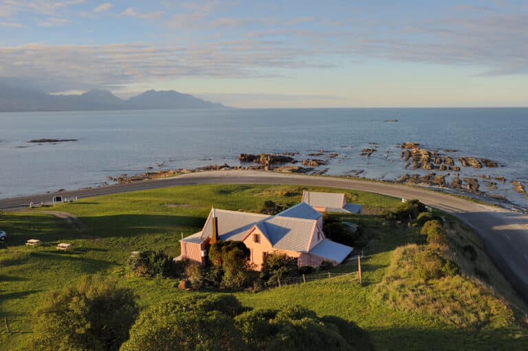 Aerial view of Fyffe House, a historic pink colonial building overlooking the ocean in Kaikōura, New Zealand, with mountains in the background.
